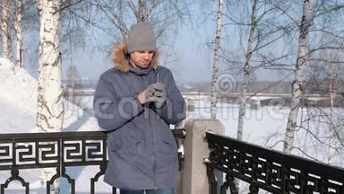 穿着带毛衣罩的蓝羽绒服的人拿起手机，在冬季公园里脱下手套。